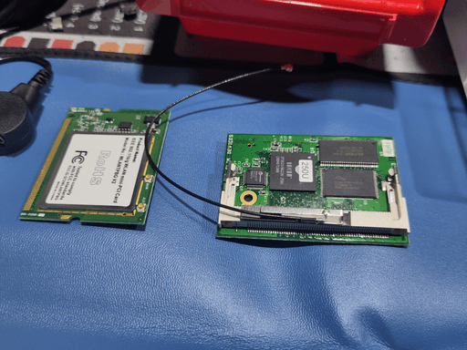 The aforementioned CPU and WIFI cards sitting side-by-side on the table. The WIFI card has a sticker declaring it as a IEEE 802.11b/g mini-PCI Card. Towards the side it has an edge-connector and most of the components are under a metal shield. The CPU card has three big chips, presumably one is the CPU, one ist the RAM and one some permanent flash for the firmware. There's also a third smaller chip and some surface mount components on the board.