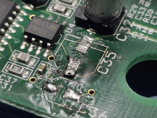 A close-up photo of the motherboard with two Capacitors removed. The footprint of the bigger one shows some damage to the solder-pads.