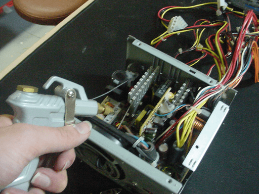 A hand holding an air gun pointed at the inside of a PC power supply.